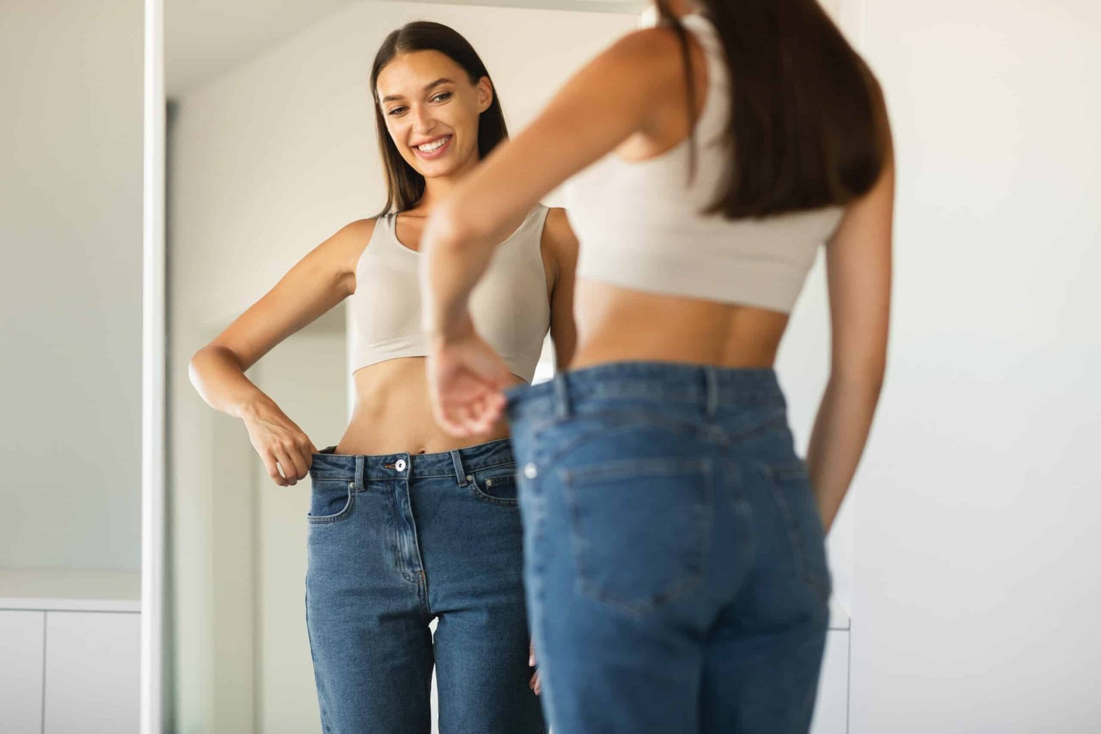 Slim Female After Successful Weight Loss Wearing Skinny Jeans Looking At Reflection In Mirror Standing At Home. Dieting And Slimming Concept. Selective Focus